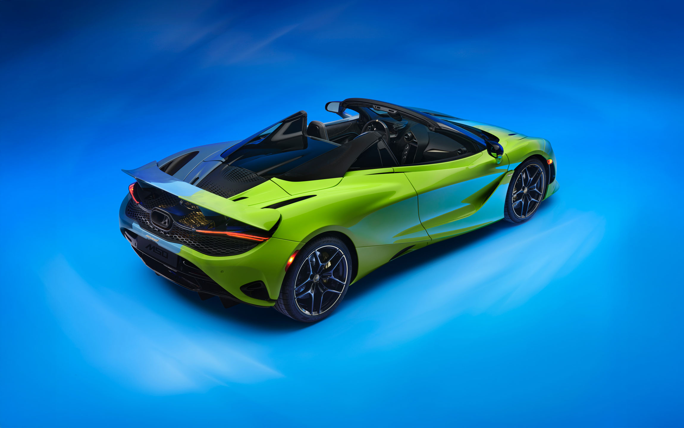 The rear side of the Project Chromology 750s by MSO and Nat Bowen, on display against a glowing blue background. The car is various overlaying shades of green and blue and can be seen parked at an angle with its headlights on.