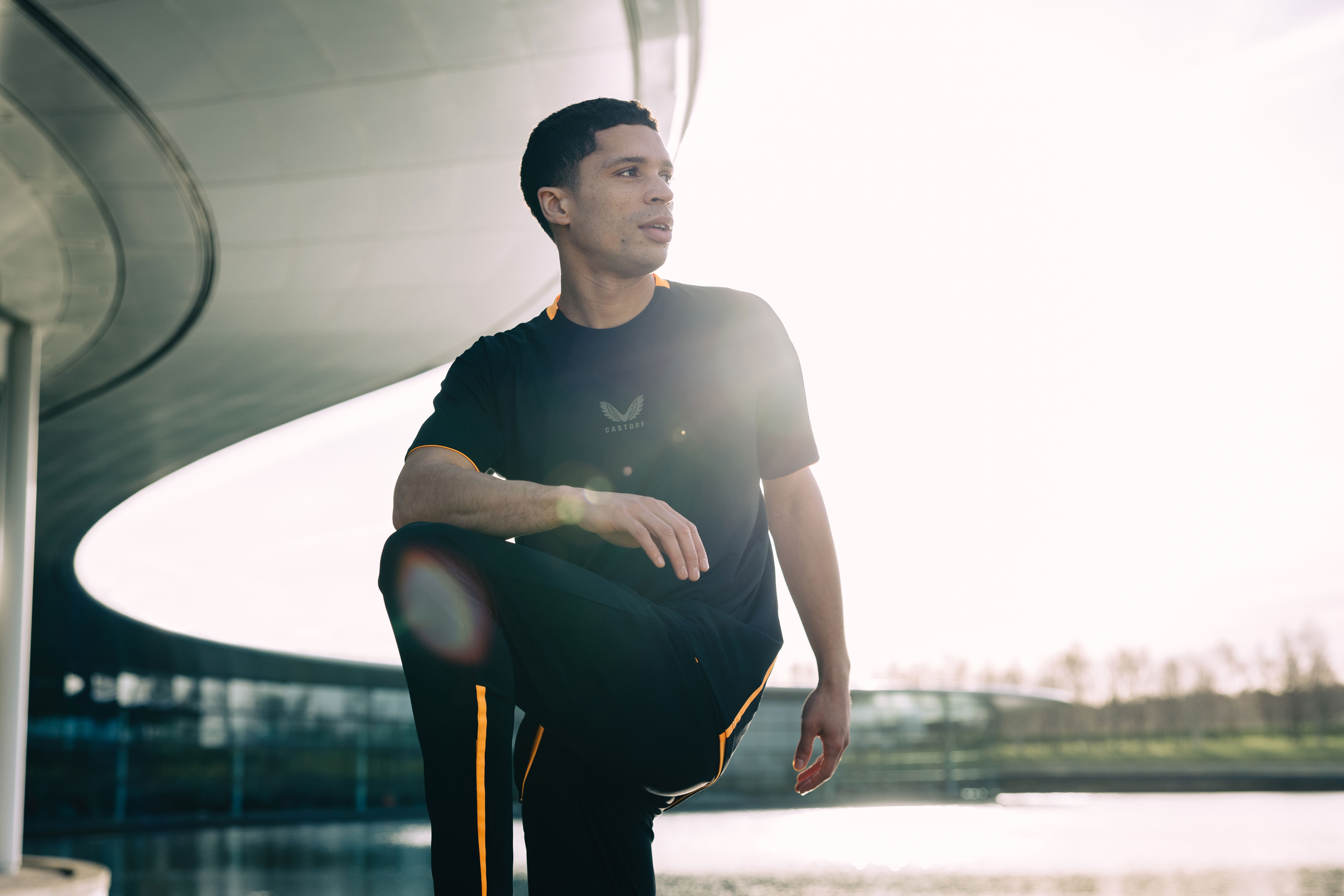 Male model wearing a black and orange Castore apparel set, stretching outside the McLaren Technology Centre at sunrise.