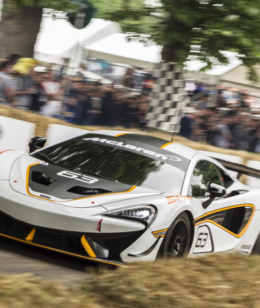 White, black and orange McLaren 570S GT4, with number 63 on the hood and side, driving around the festival of speed track at speed. A blurry crowd of people around the track can be seen in the background.