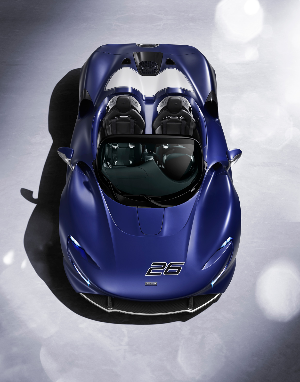 An aerial top-down view of a blue McLaren Elva roadster with a white racing stripe and the number 26 on the hood, showcasing its unique open cockpit and streamlined body.