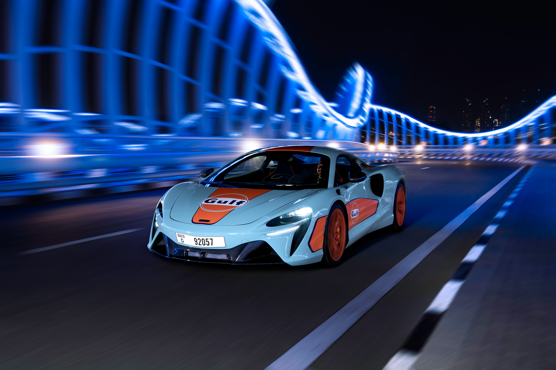 Gulf Branded McLaren driving over a lit up city bridge at night.