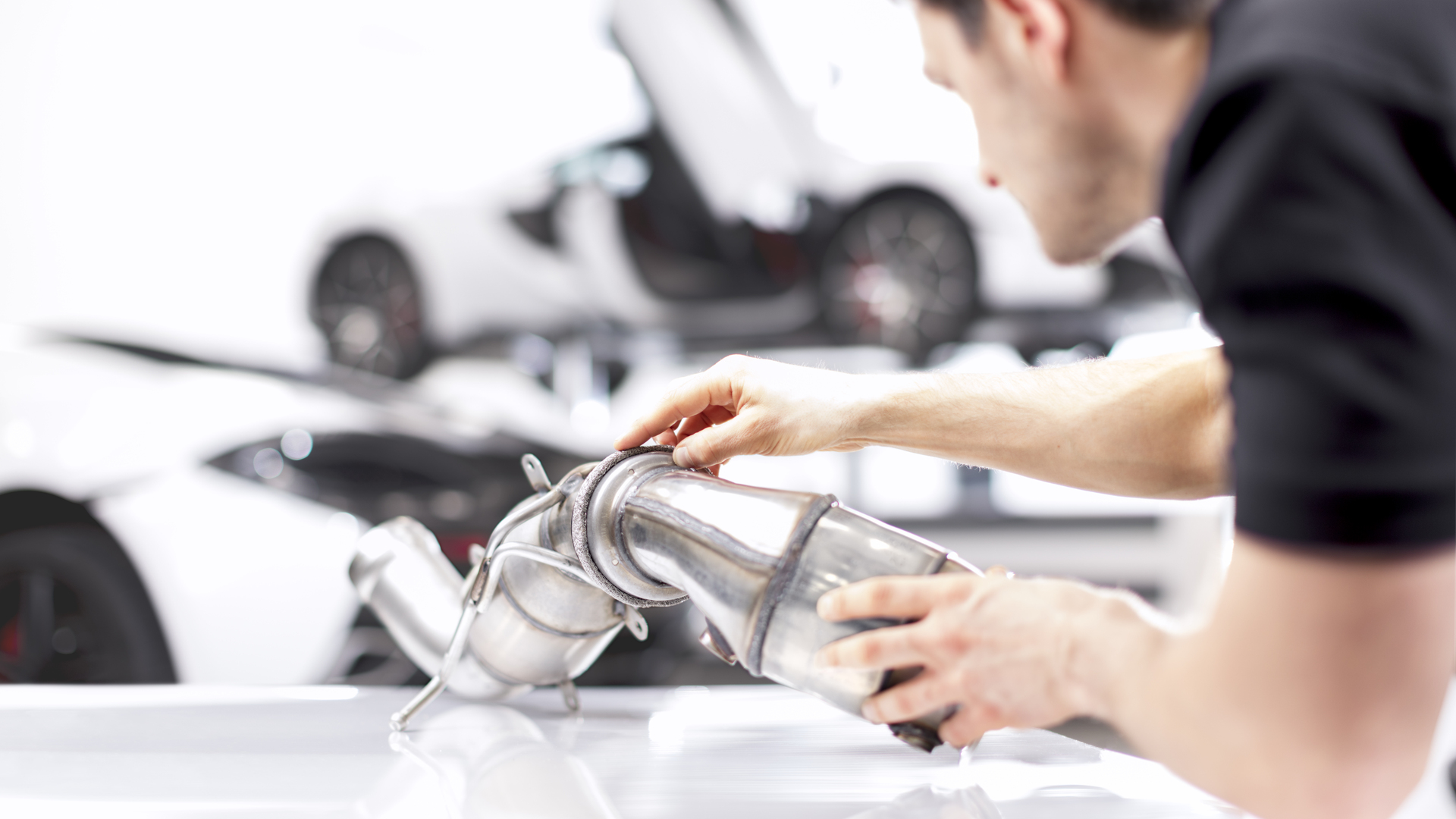 A McLaren technician carefully inspecting a car exhaust system in a workshop, with a supercar in the background.