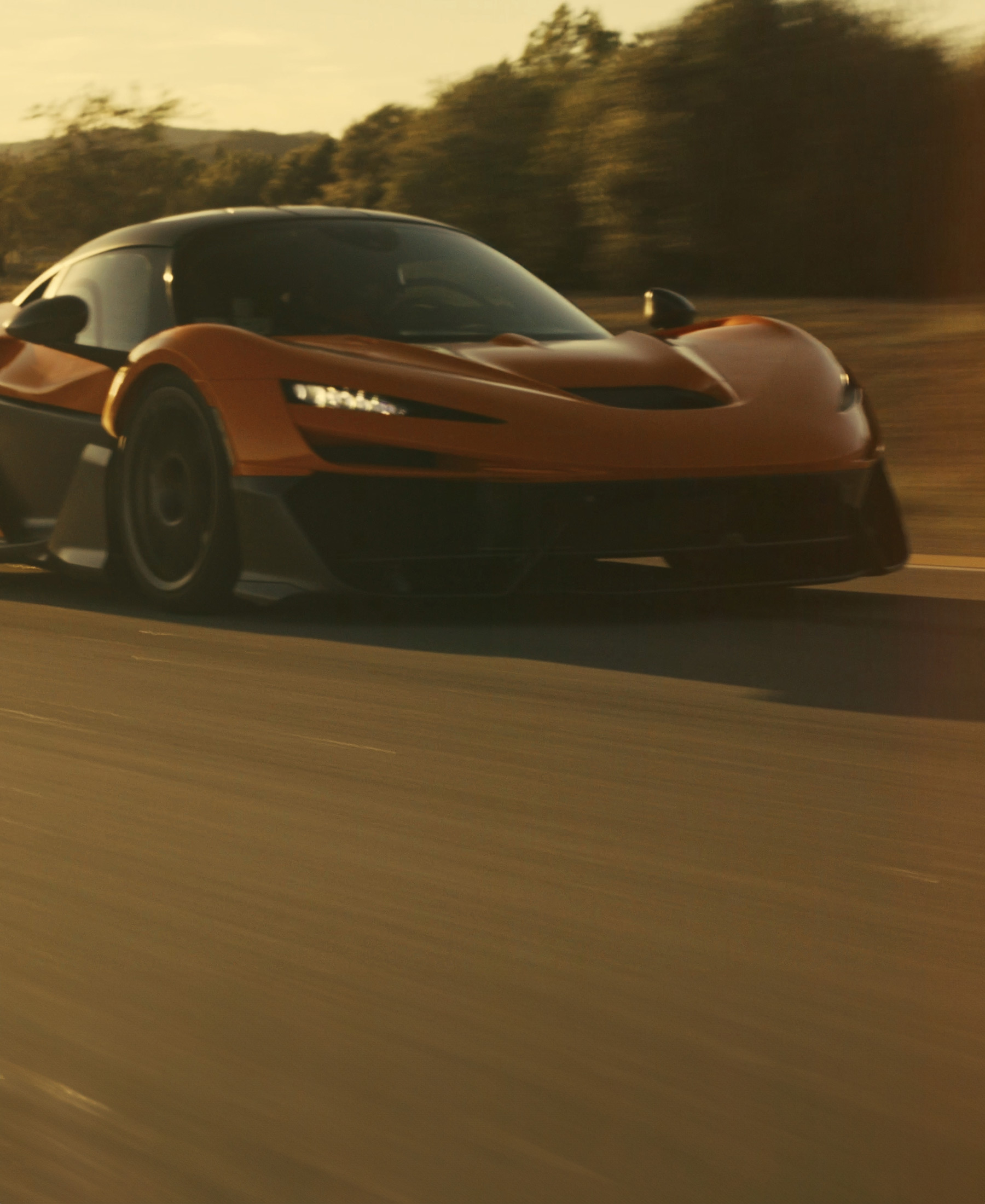 A dynamic, low-angle shot of an orange and black McLaren W1 with its headlights on, speeding down a road under a warm, sunlit sky, with blurred trees in the background conveying motion.