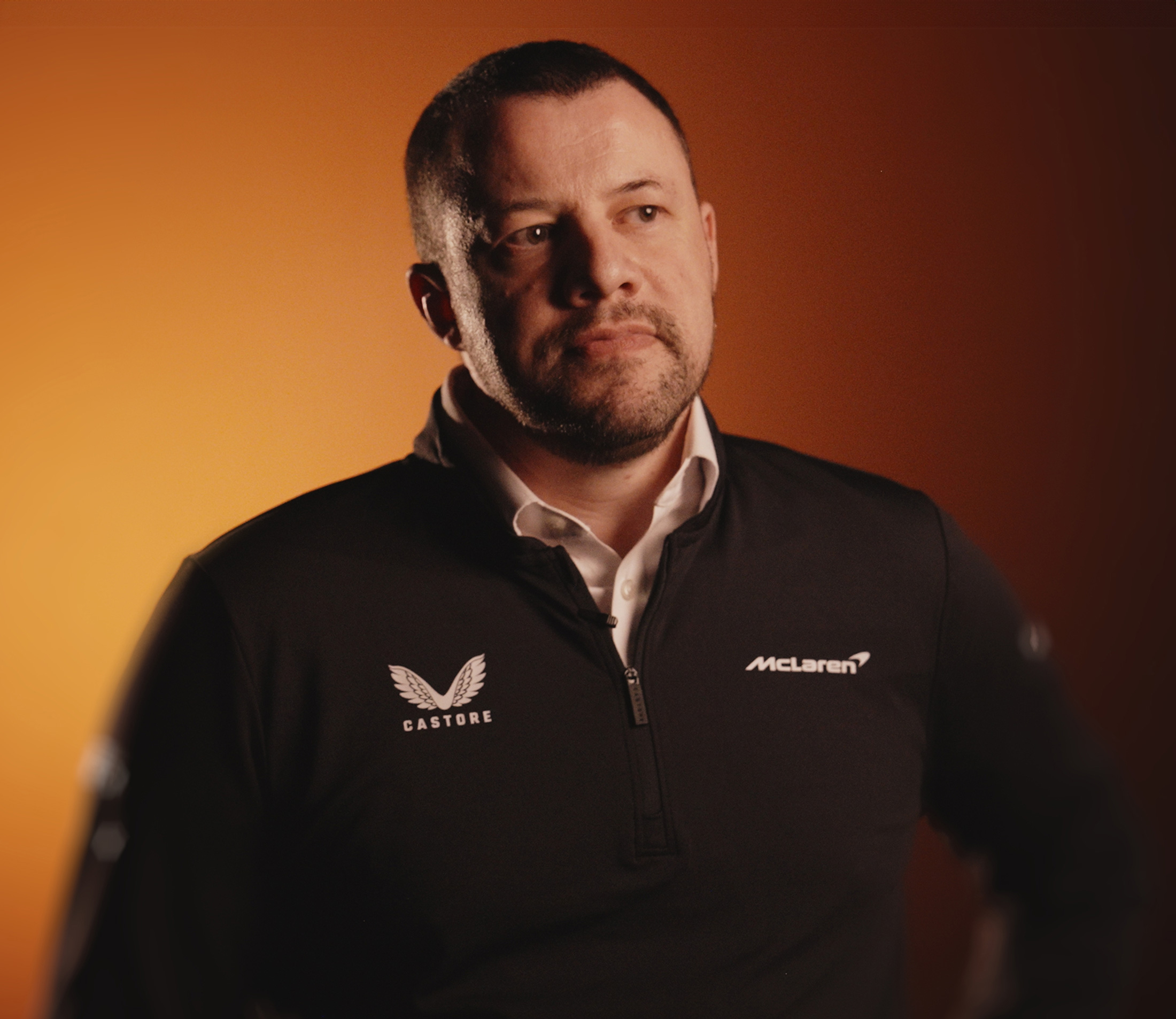 A man with short hair and a beard, wearing a black McLaren-branded jacket, looks thoughtfully to his right against an orange background.