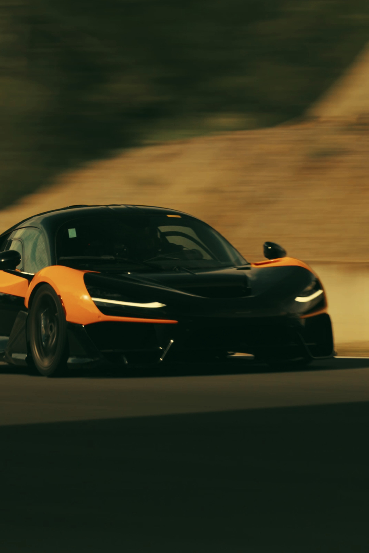 A dynamic side-on shot of an orange and black McLaren W1, showcasing its headlights and livery as it speeds along a track with a blurred background.