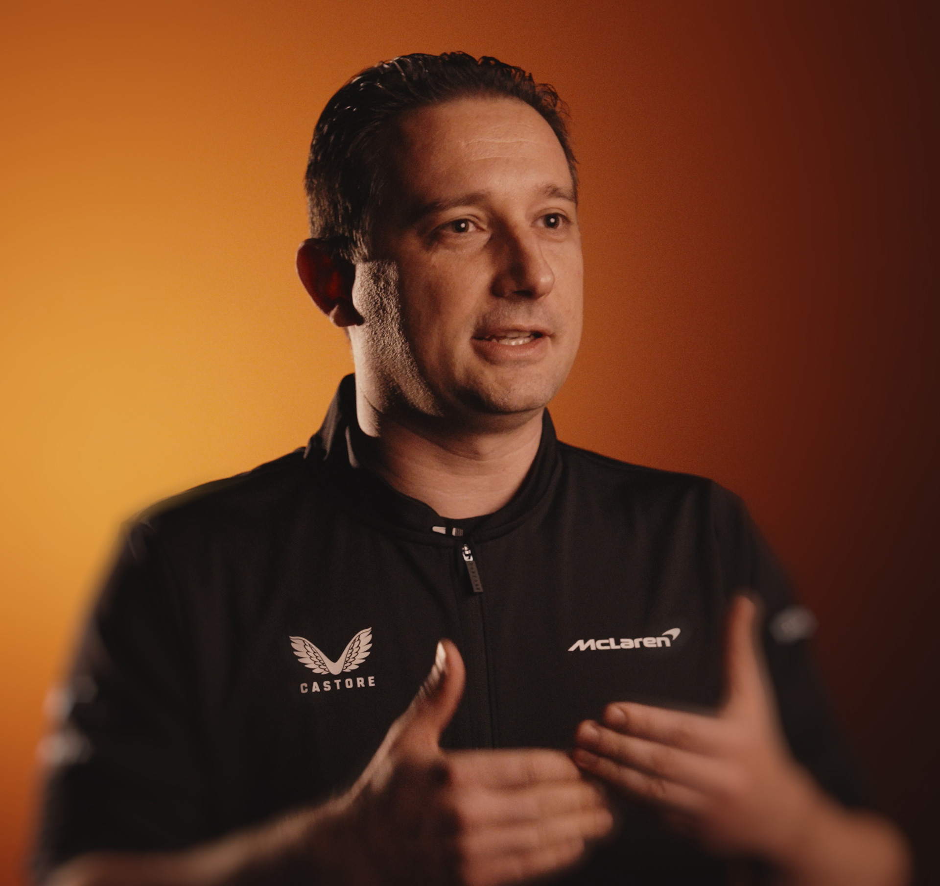 A man in a black jacket with McLaren and Castore logos gestures with his hands while speaking, against a warm orange background.