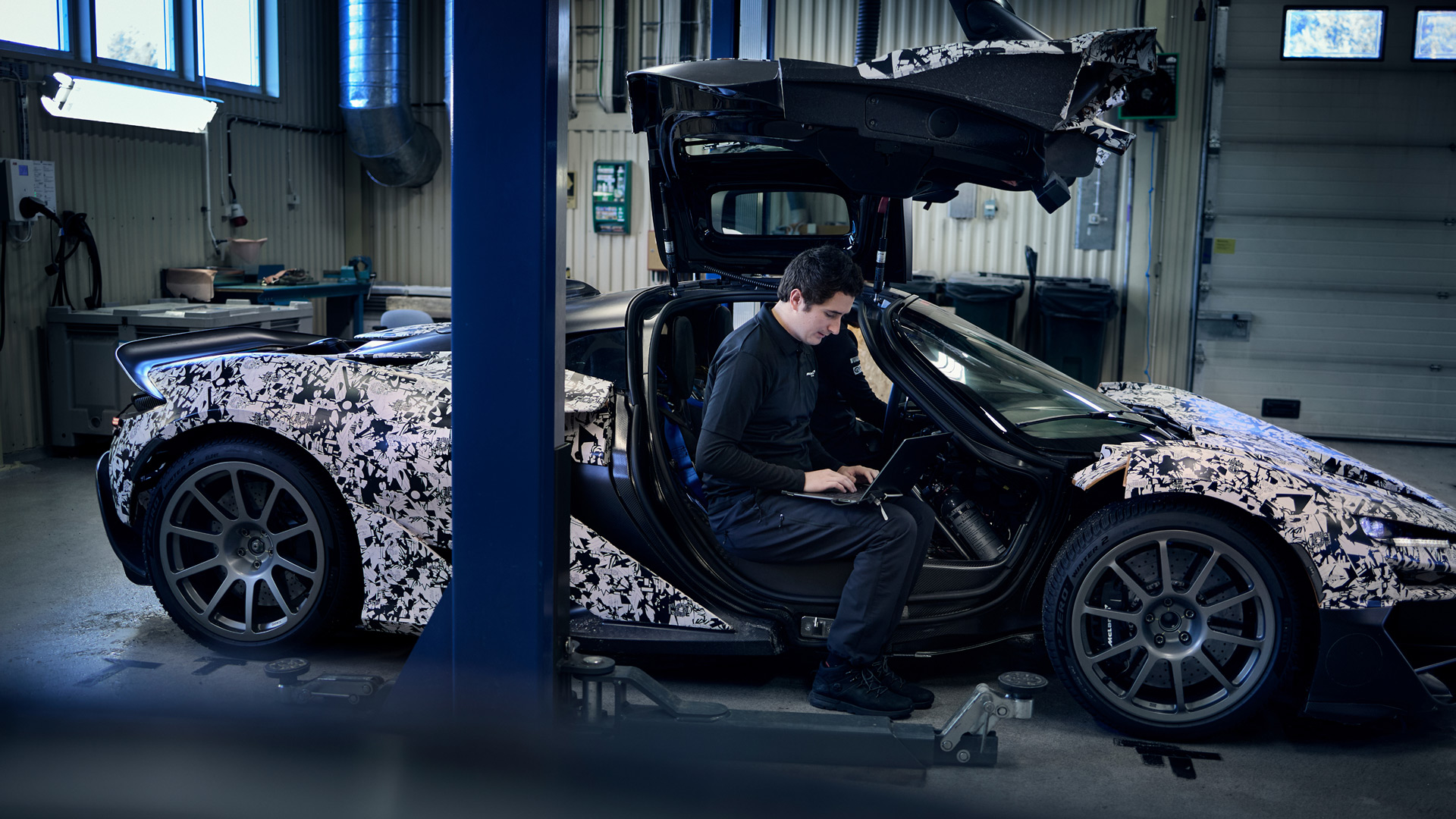 A technician is seated inside a camouflage-wrapped McLaren W1 prototype, which has its door open and is on a lift in a garage, working on a laptop.