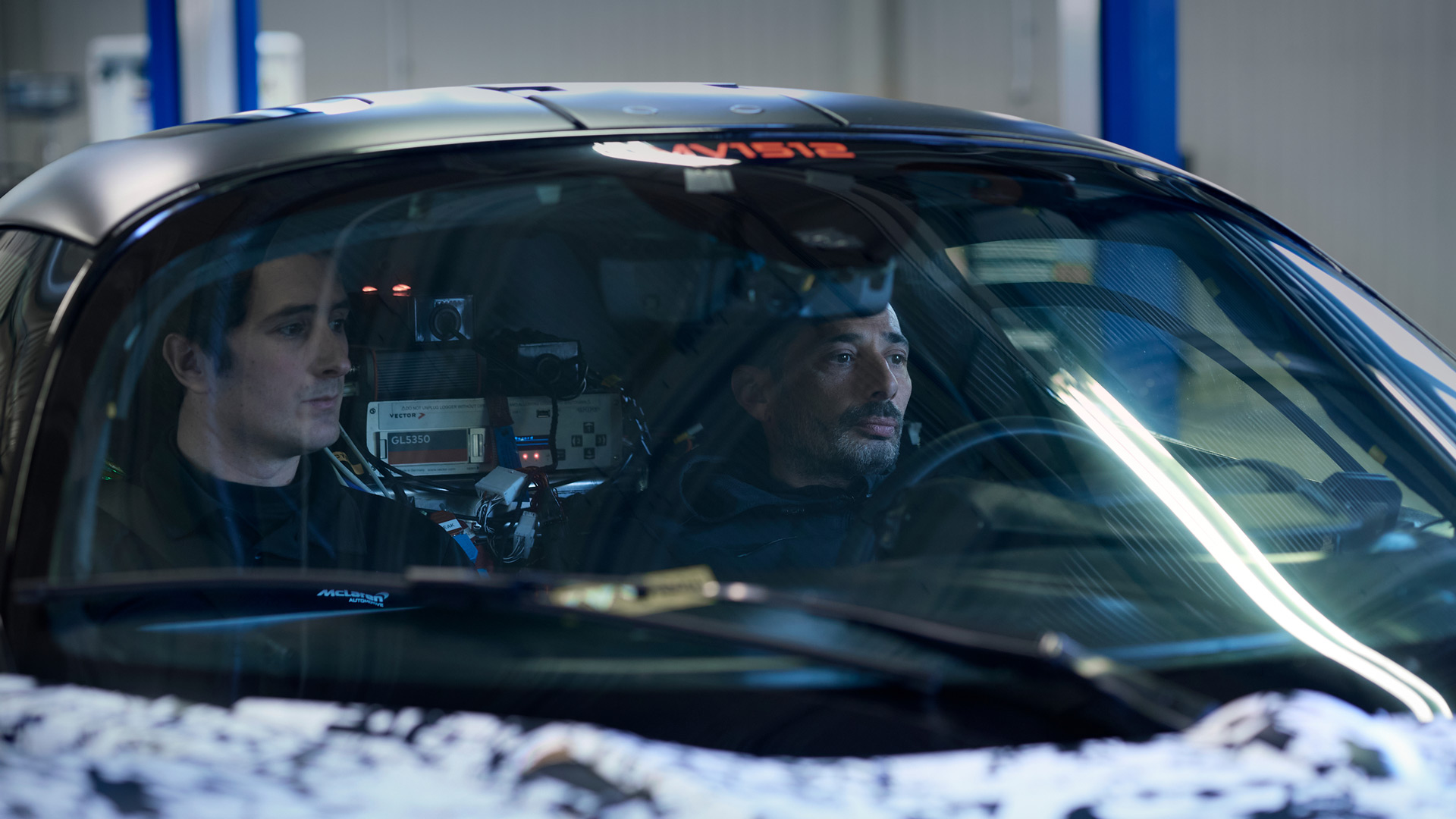 Two men are visible through the windshield of a McLaren W1, which is parked indoors with a testing rig visible inside.