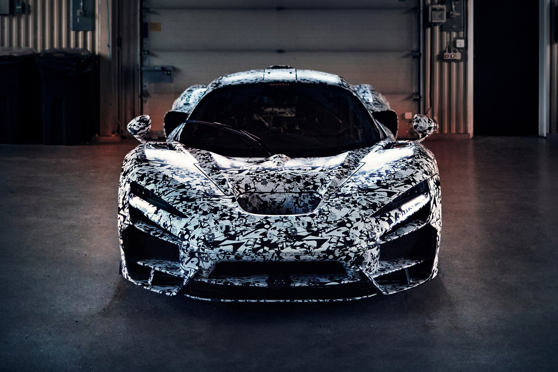 A front view of a McLaren W1 prototype in a black and white camouflage wrap, parked inside what appears to be a garage, with its headlights illuminated.