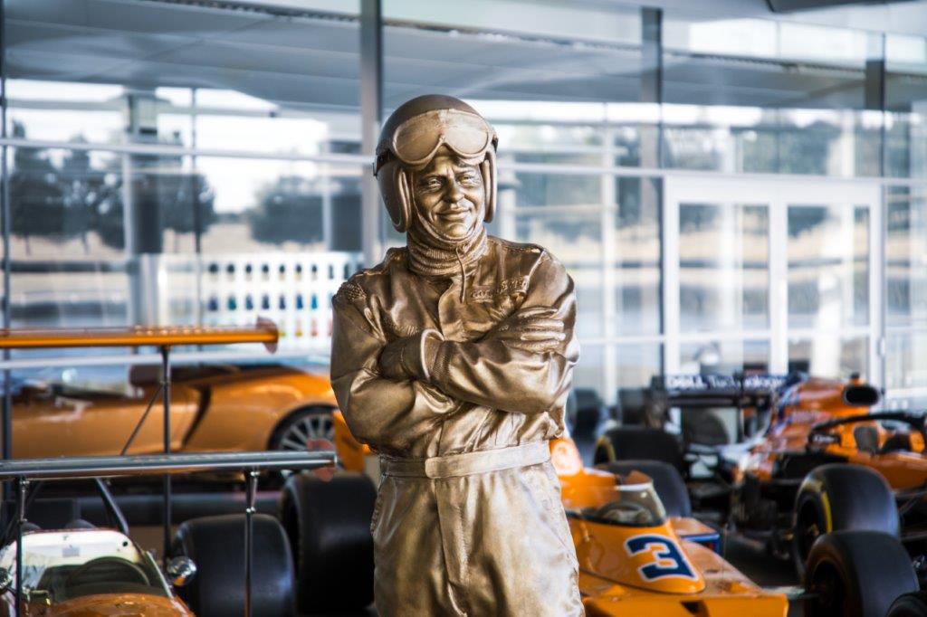 A bronze statue of racing legend Bruce McLaren, founder of McLaren, standing with his arms crossed.